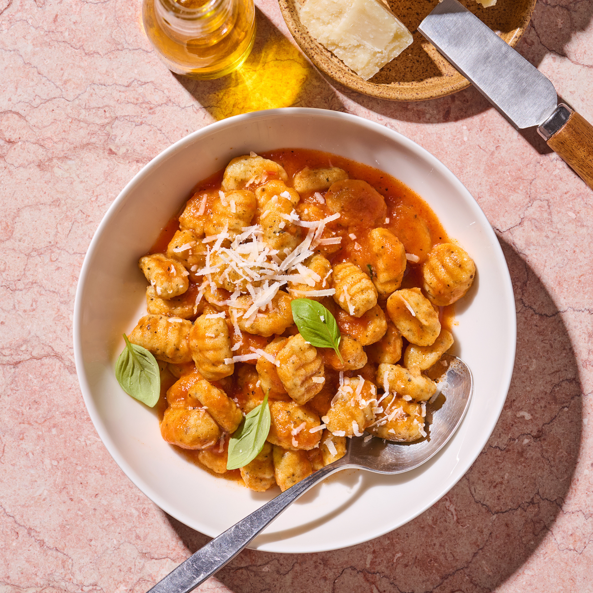 Weiße Schüssel mit Gnocchi in Tomatensauce, garniert mit Basilikum und geriebenem Käse, daneben Olivenöl und Käse.