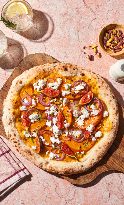 Pizza mit Feta, roten Zwiebeln und Feigen auf Holzbrett, daneben Schälchen mit Pistazien und zwei Gläsern Wasser.