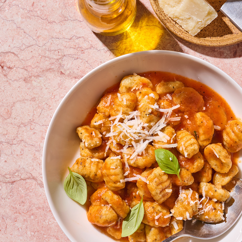 Ein Teller Gnocchi mit Tomatensauce, garniert mit Basilikum und geriebenem Käse, daneben ein Glas Olivenöl.