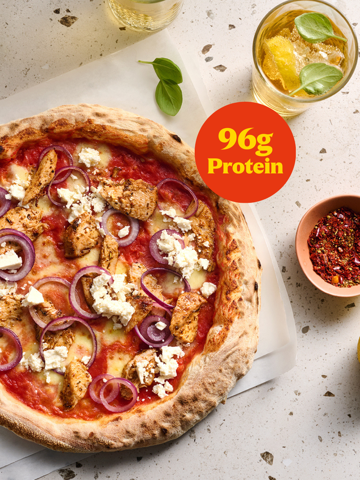 Pizza mit Hähnchen, roten Zwiebeln und Feta, umgeben von Getränken und Kräutern, Text: "96g Protein".