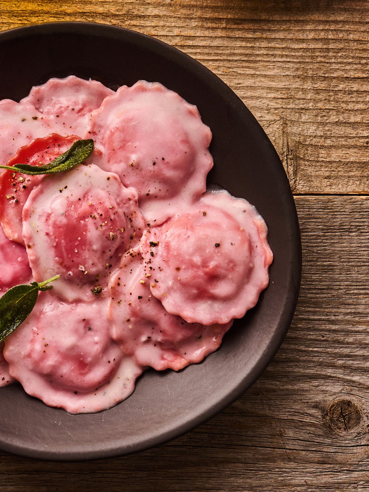 Rosa Ravioli auf einem dunklen Teller, garniert mit Salbeiblättern und Pfeffer auf einem Holztisch.