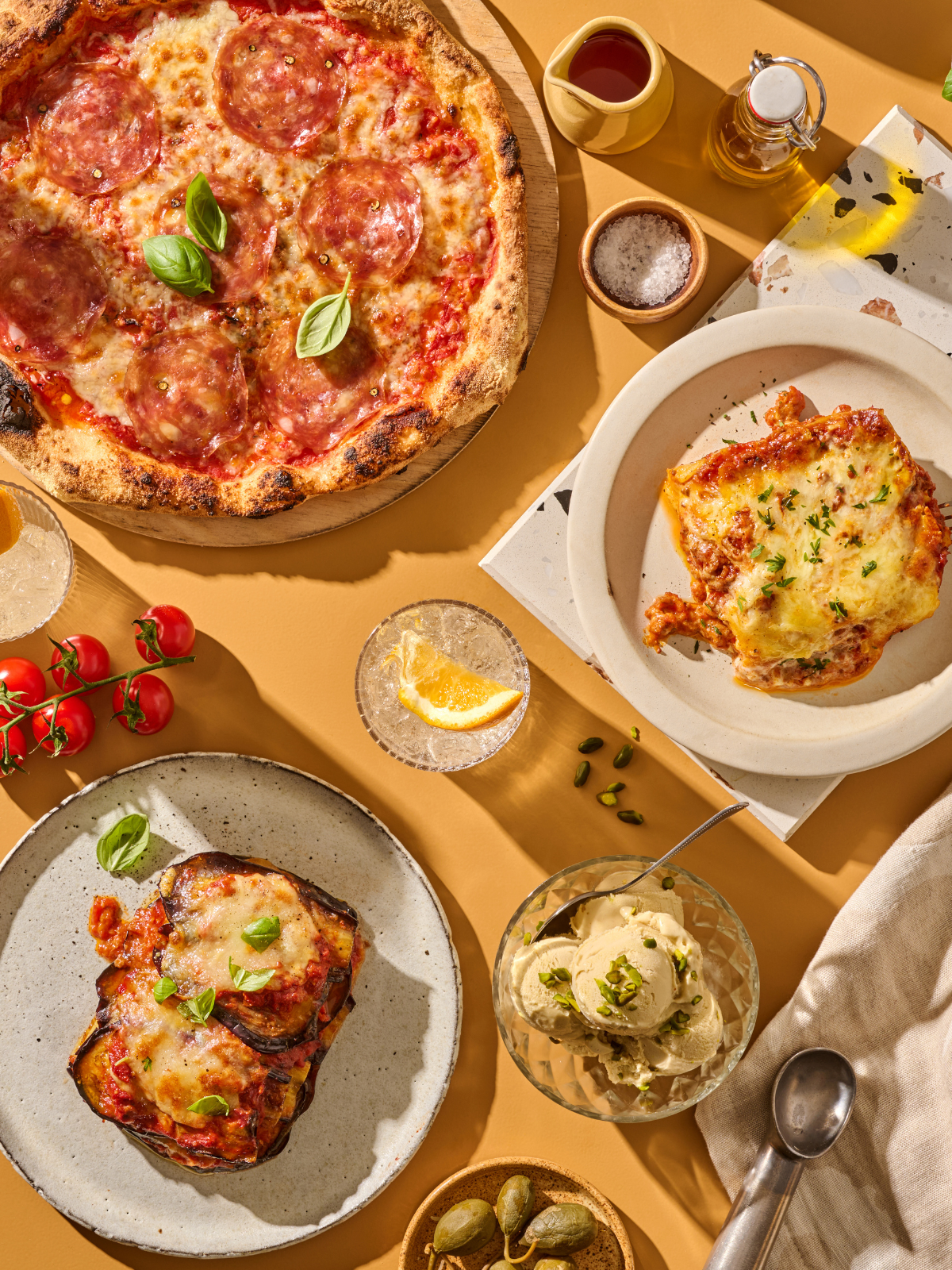 Pizza, Lasagne, Auberginenauflauf und Pistazieneis auf einem Tisch mit Getränken und Kirschtomaten.