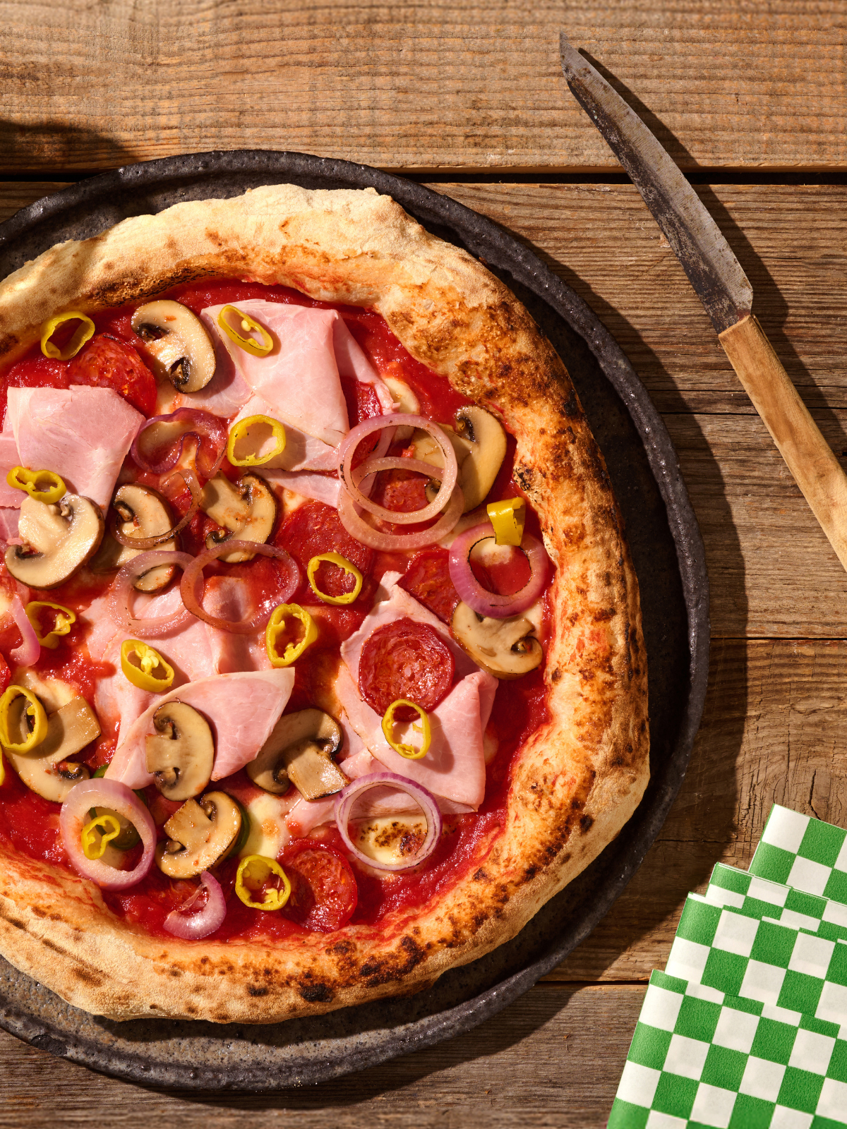 Pizza mit Schinken, Pilzen, Peperoni und Zwiebeln auf einem Tisch mit einem Messer und grün-weißen Servietten.