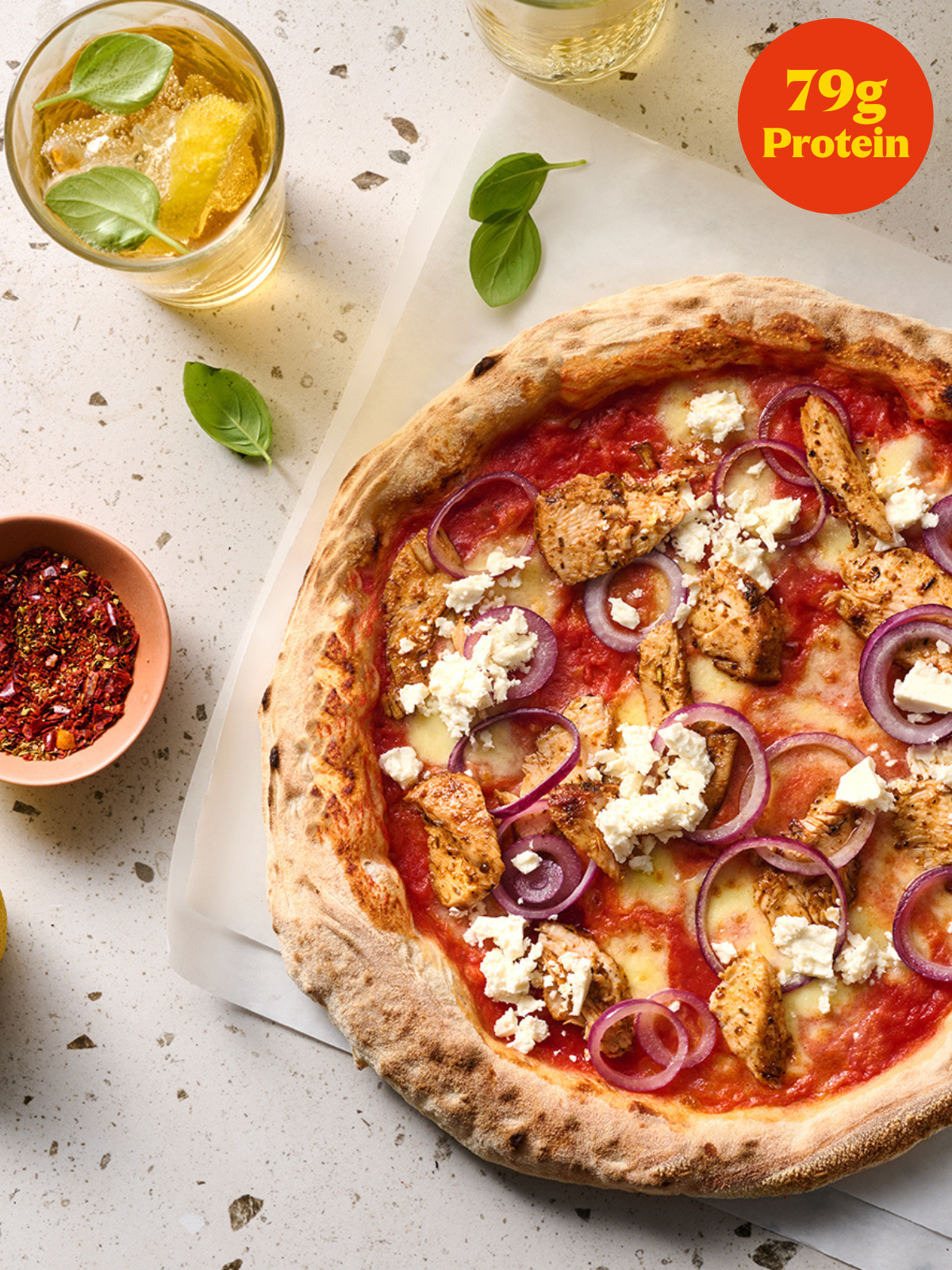 Pizza mit Hähnchen, roten Zwiebeln und Feta auf einem Tisch mit Getränken und Kräutern, zeigt "79g Protein".