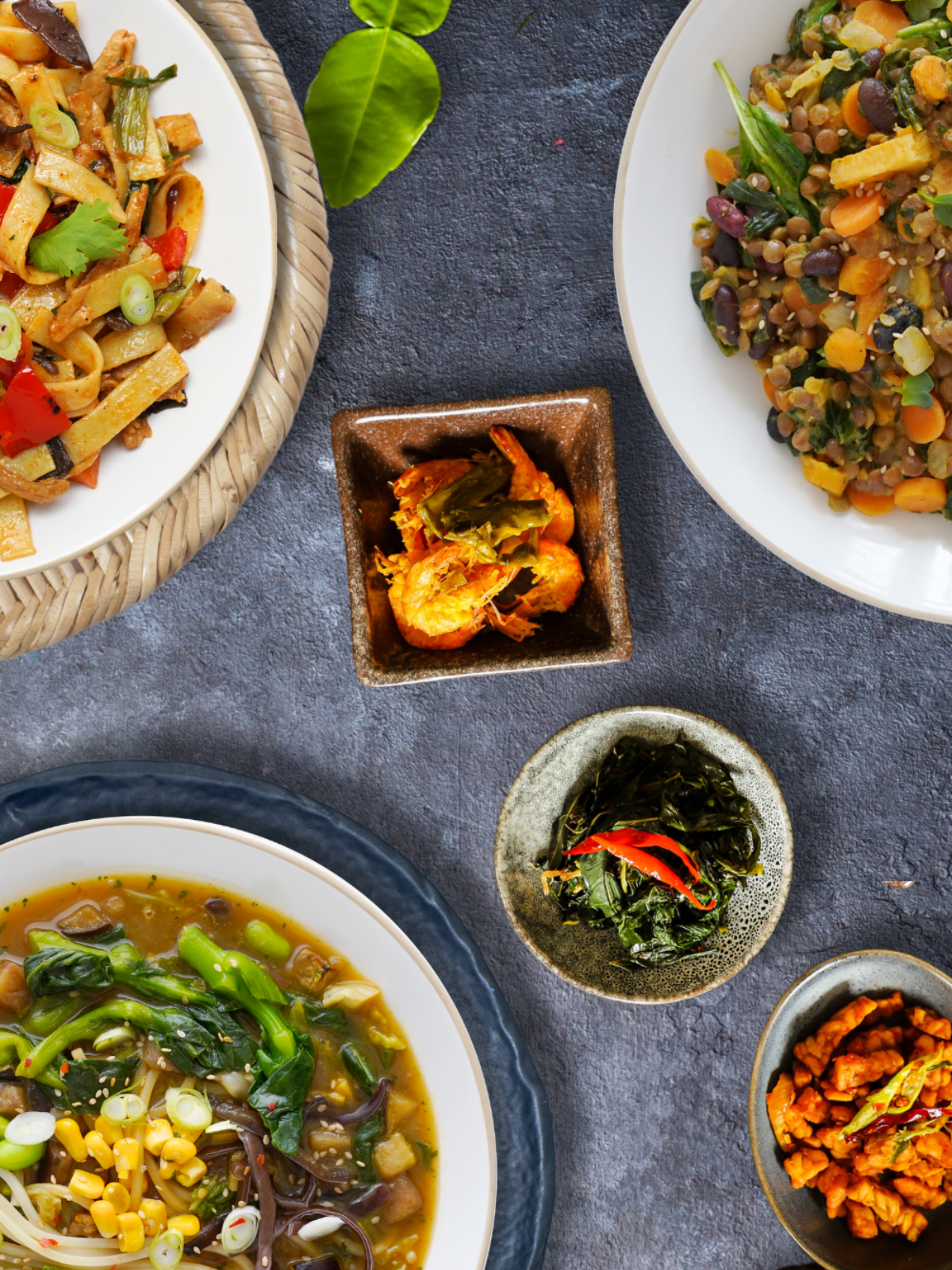 Eine Auswahl an bunten asiatischen Gerichten auf runden Tellern, flach auf einem dunklen Tisch angeordnet.
