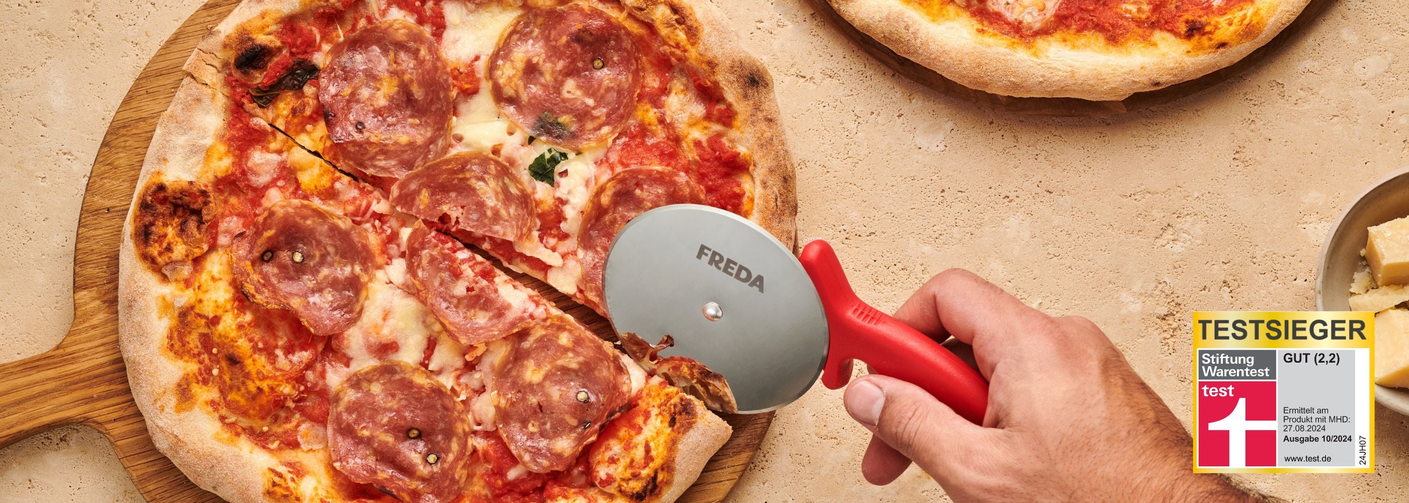 Eine Hand schneidet Pizza mit einem Pizzaschneider. Daneben liegt Parmesan und ein Testsiegel von Stiftung Warentest.
