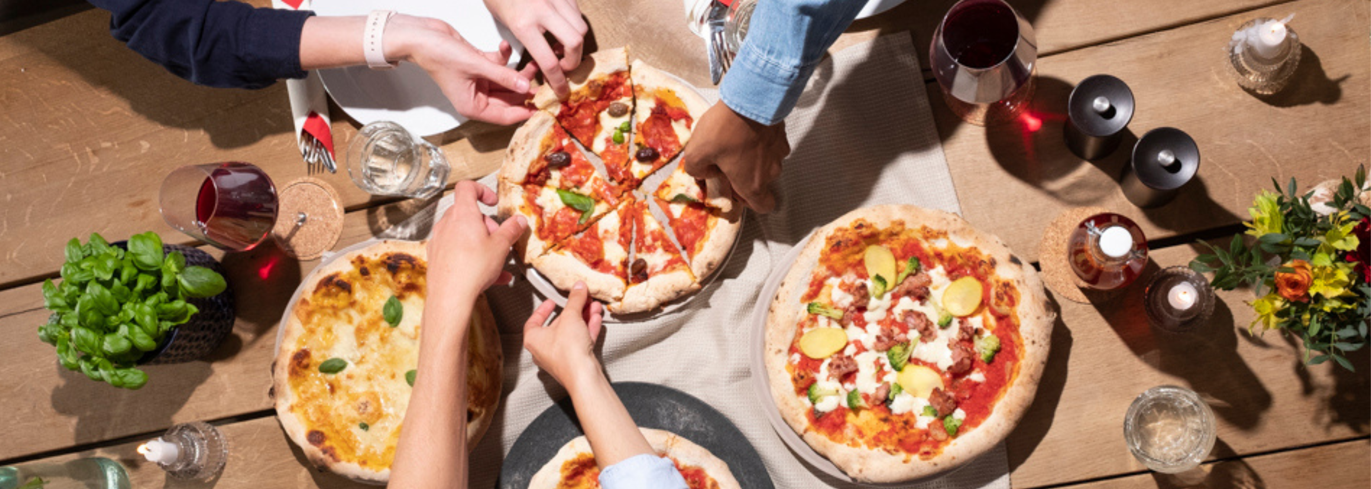 Mehrere Personen greifen nach Pizzastücken auf einem Holztisch mit Gläsern, Besteck und Dekoration.