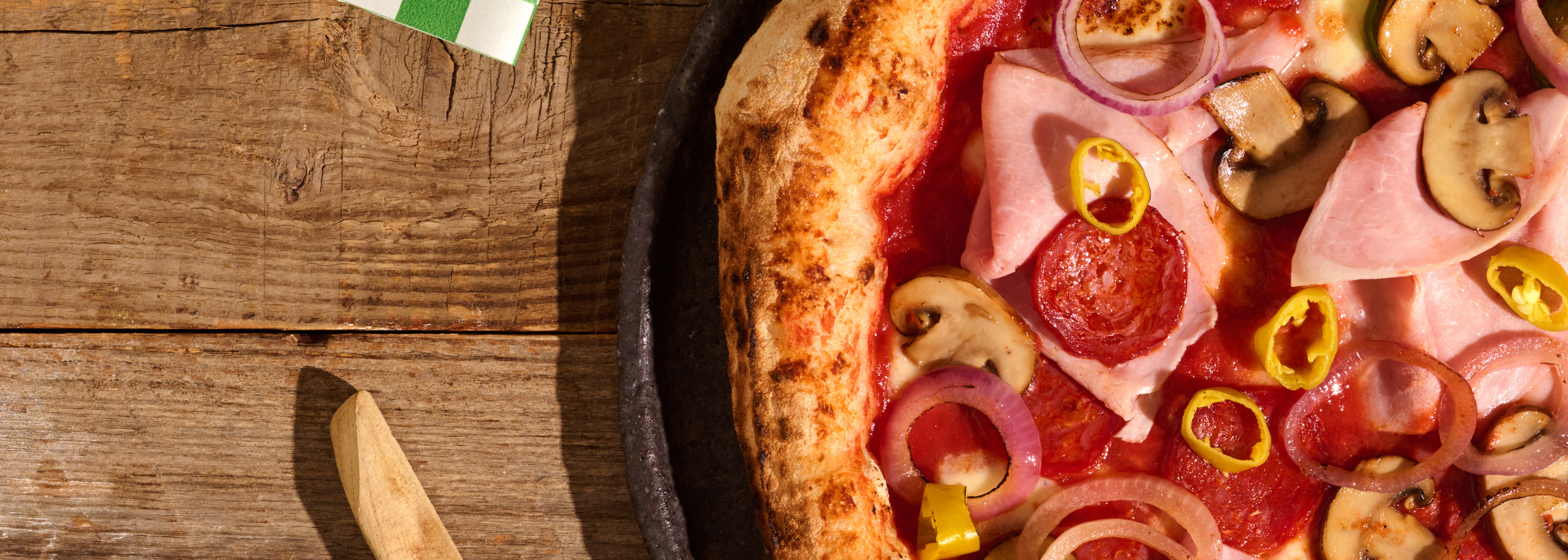 Nahaufnahme einer Pizza mit Peperoni, Schinken, Pilzen, roten Zwiebeln und gelben Peperoni auf einem Holztisch.