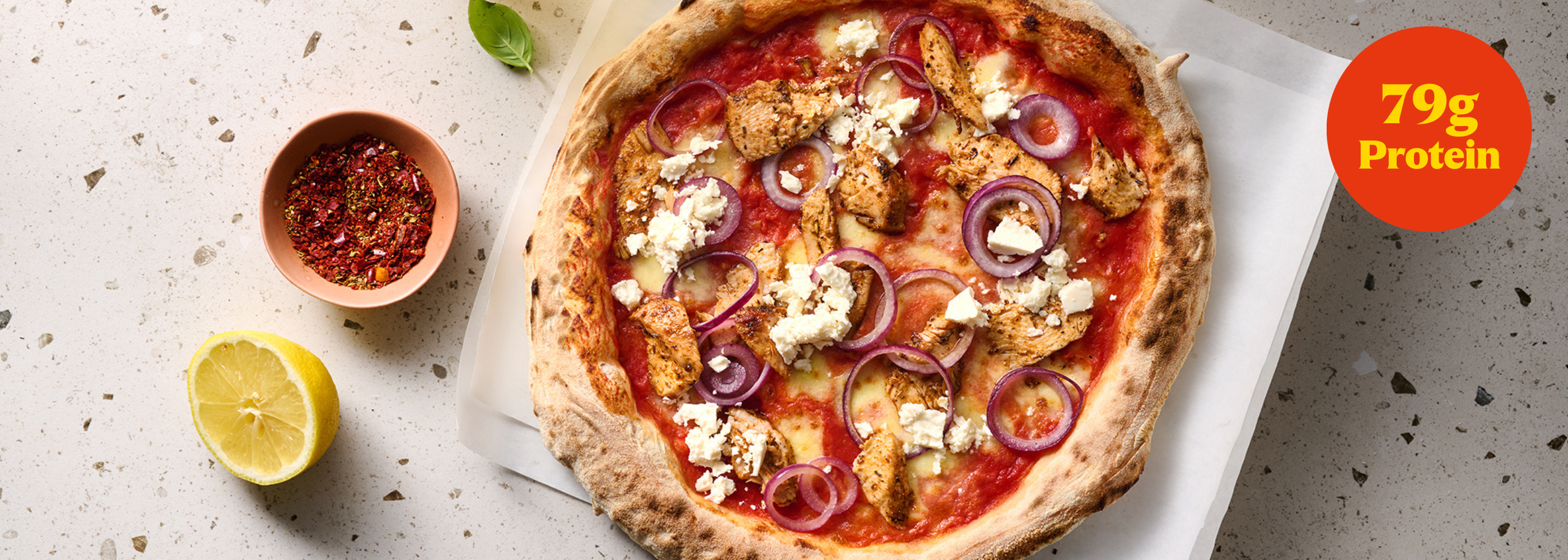 Pizza mit gebratenem Hähnchen, roten Zwiebeln und Feta, daneben eine halbe Zitrone und Gewürze. Text: "79g Protein".