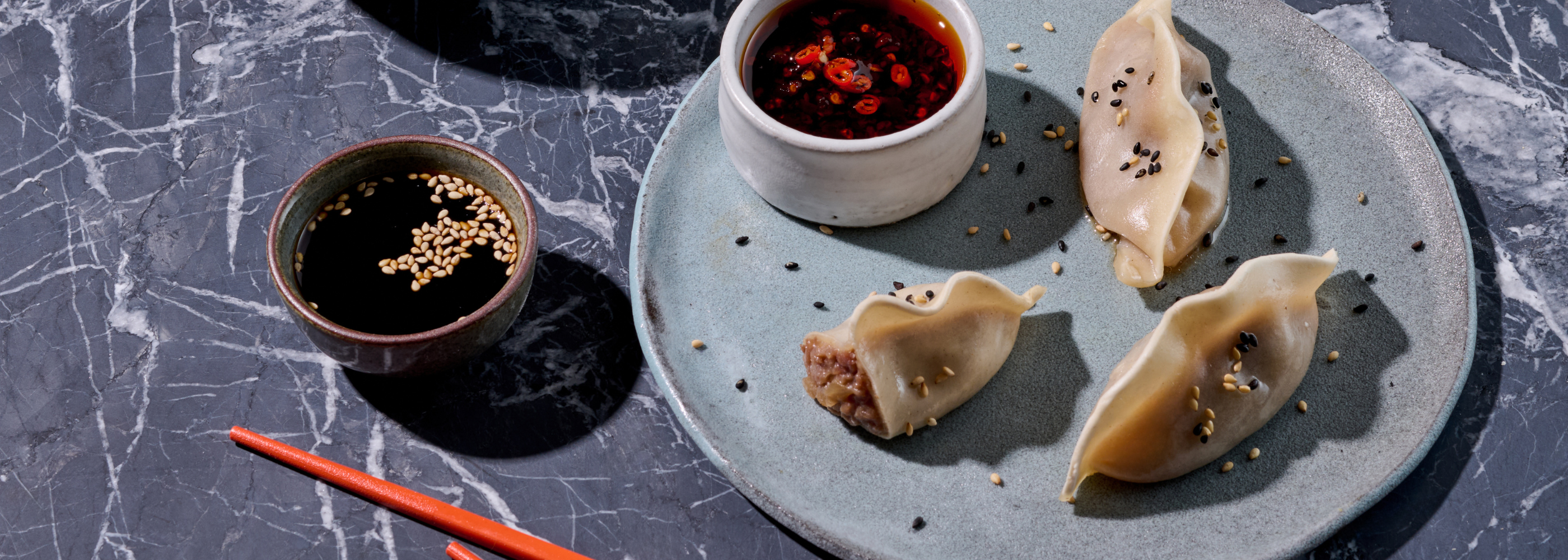 Drei Dumplings mit Sesam auf einem Teller neben zwei Schälchen mit Dips auf einer Marmorplatte.