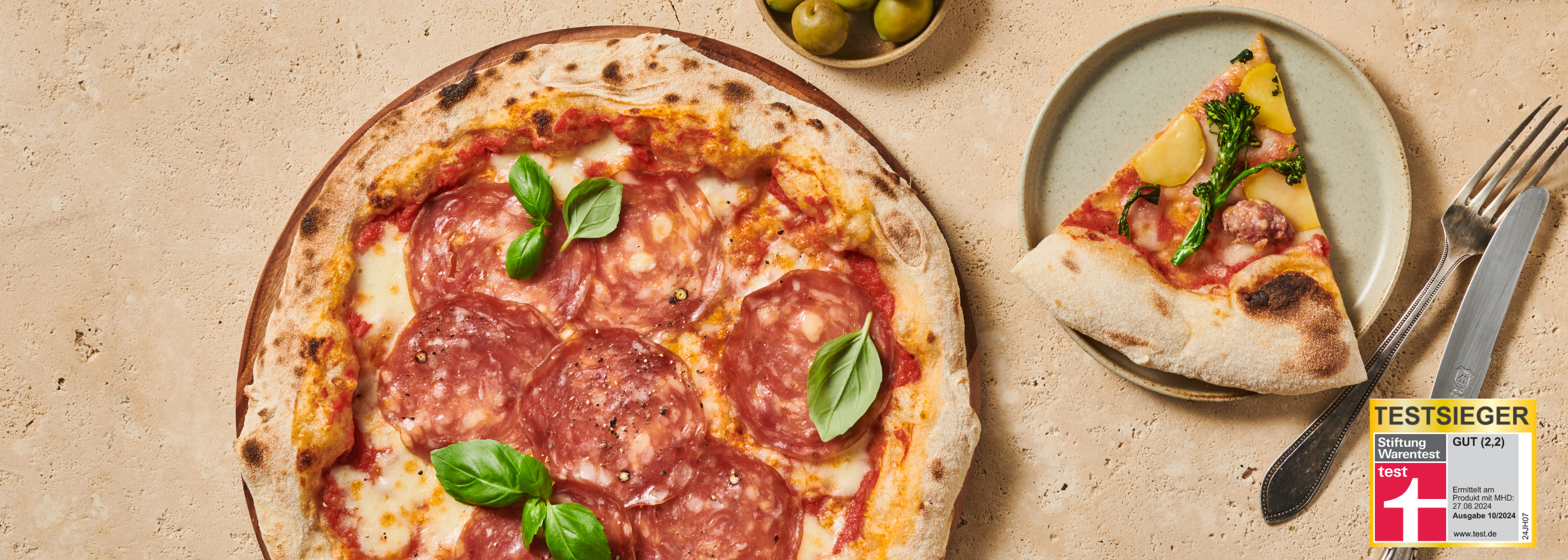 Pizza mit Salami, Basilikum und eine Scheibe Kartoffelpizza auf Teller, daneben Besteck und ein Testsieger-Siegel.