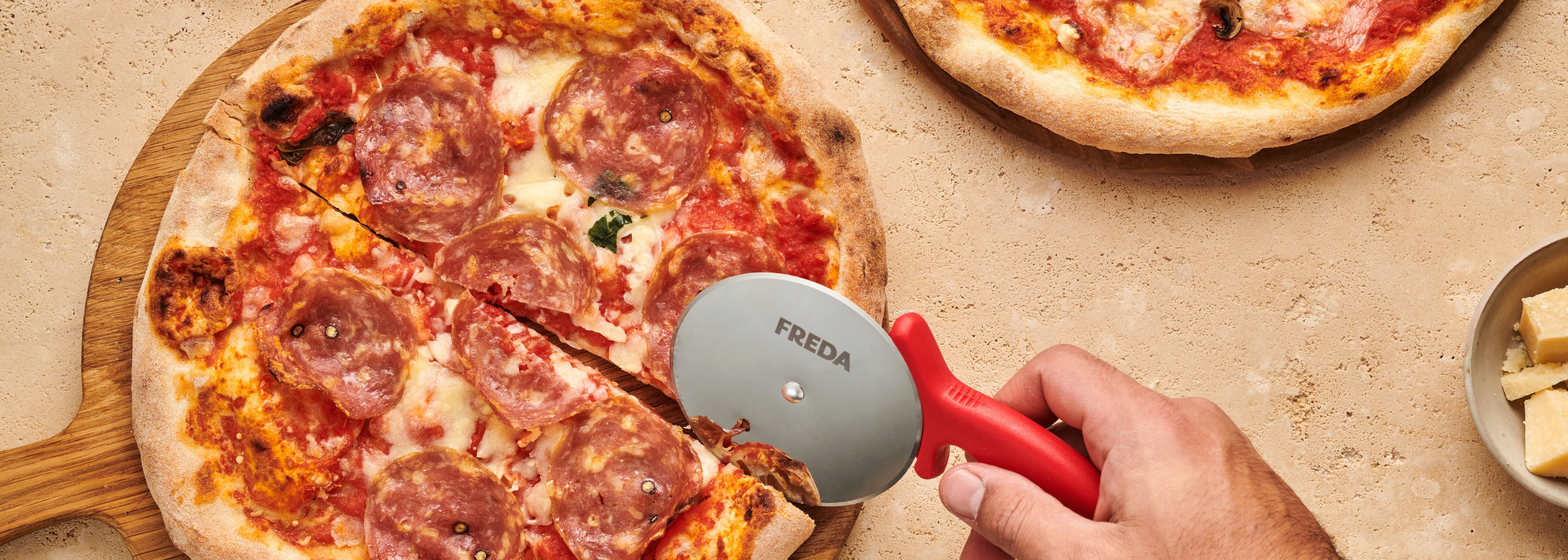 Hand schneidet Salami-Pizza mit einem Pizzaschneider auf einem hölzernen Pizzabrett.