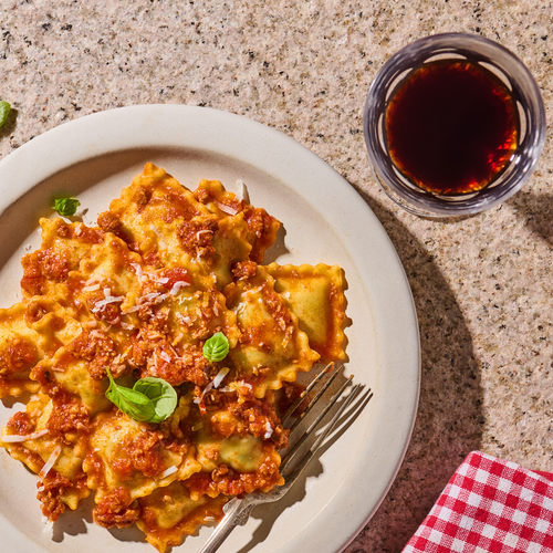 Ein Teller Ravioli mit Tomatensauce, Basilikum und Parmesan, daneben ein Glas Rotwein auf einem Steinuntergrund.
