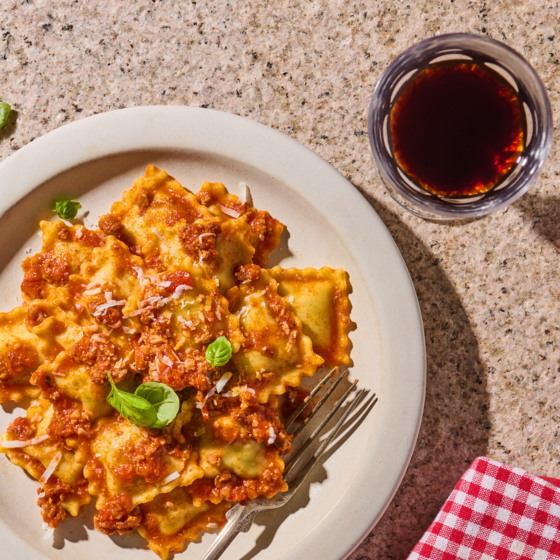Ein Teller Ravioli mit Tomatensauce, Basilikum und Parmesan, daneben ein Glas Rotwein auf einem Steinuntergrund.
