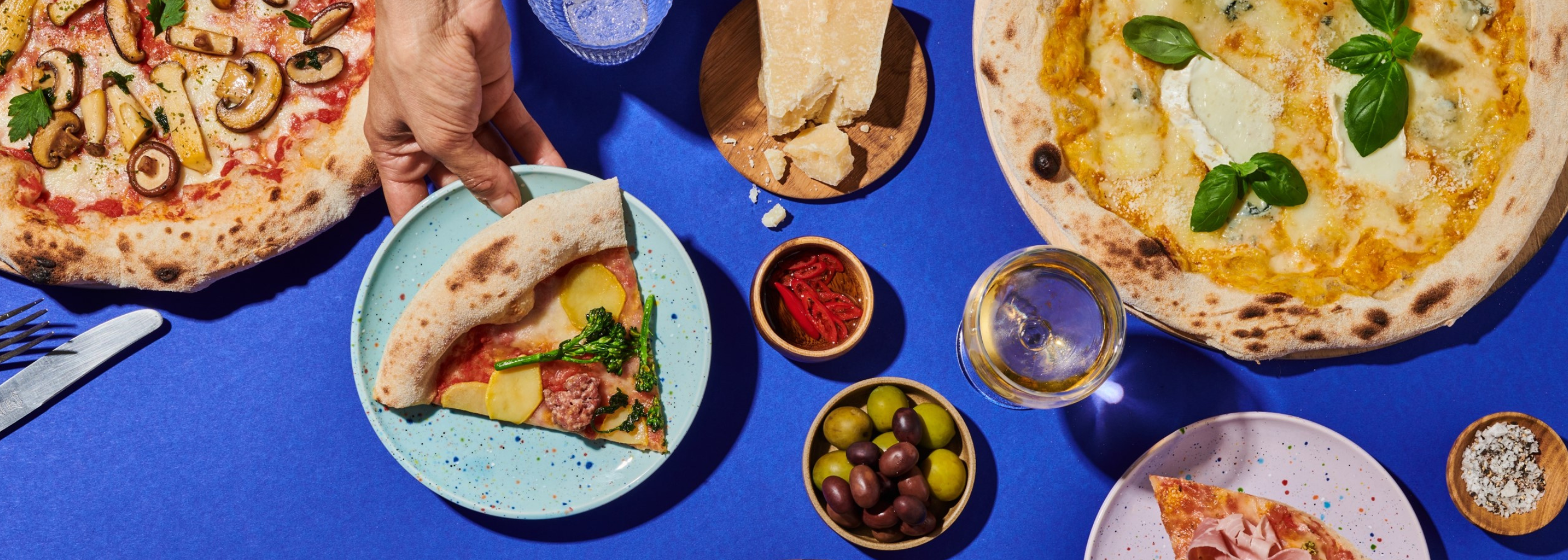 Verschiedene Pizzen auf blauem Tisch, mit Oliven, Parmesan, Prosecco und einer Hand, die ein Stück aufnimmt.
