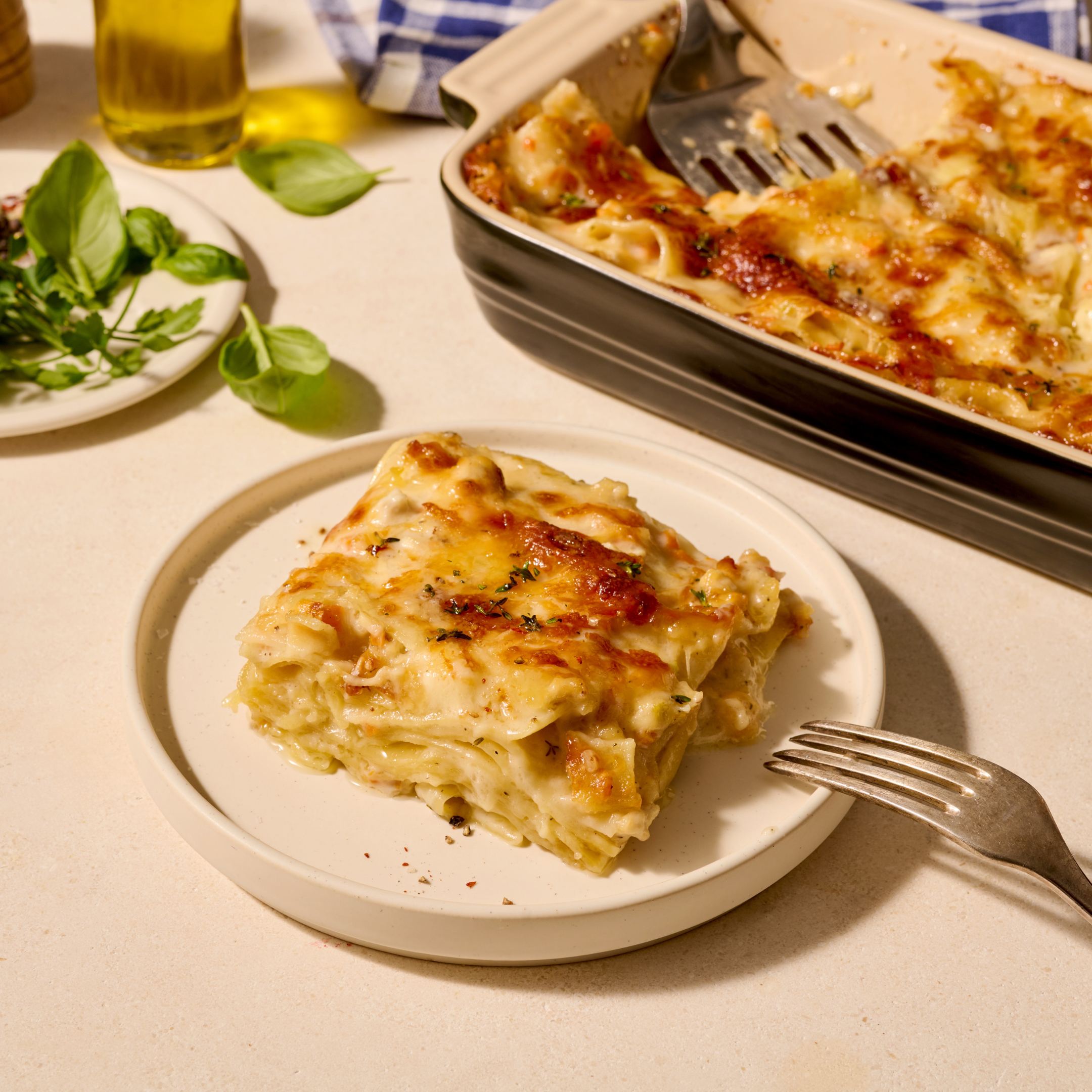 Ein Stück Lasagne auf einem Teller neben einer Auflaufform mit mehr Lasagne und frischen Basilikumblättern.