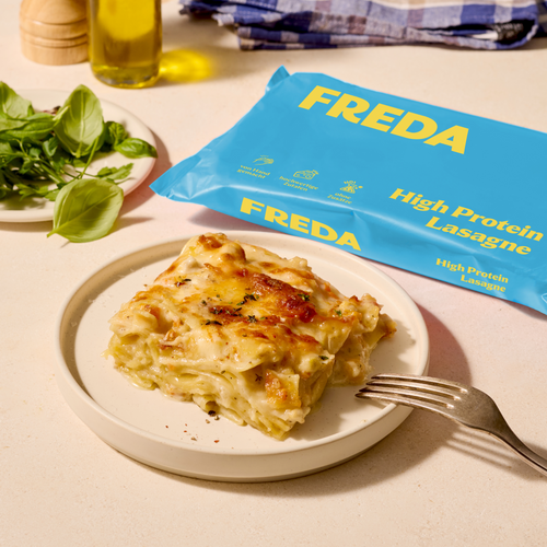 Ein Teller mit einer Portion Lasagne, daneben eine Packung "FREDA High Protein Lasagne" und ein Teller mit Basilikumblättern.