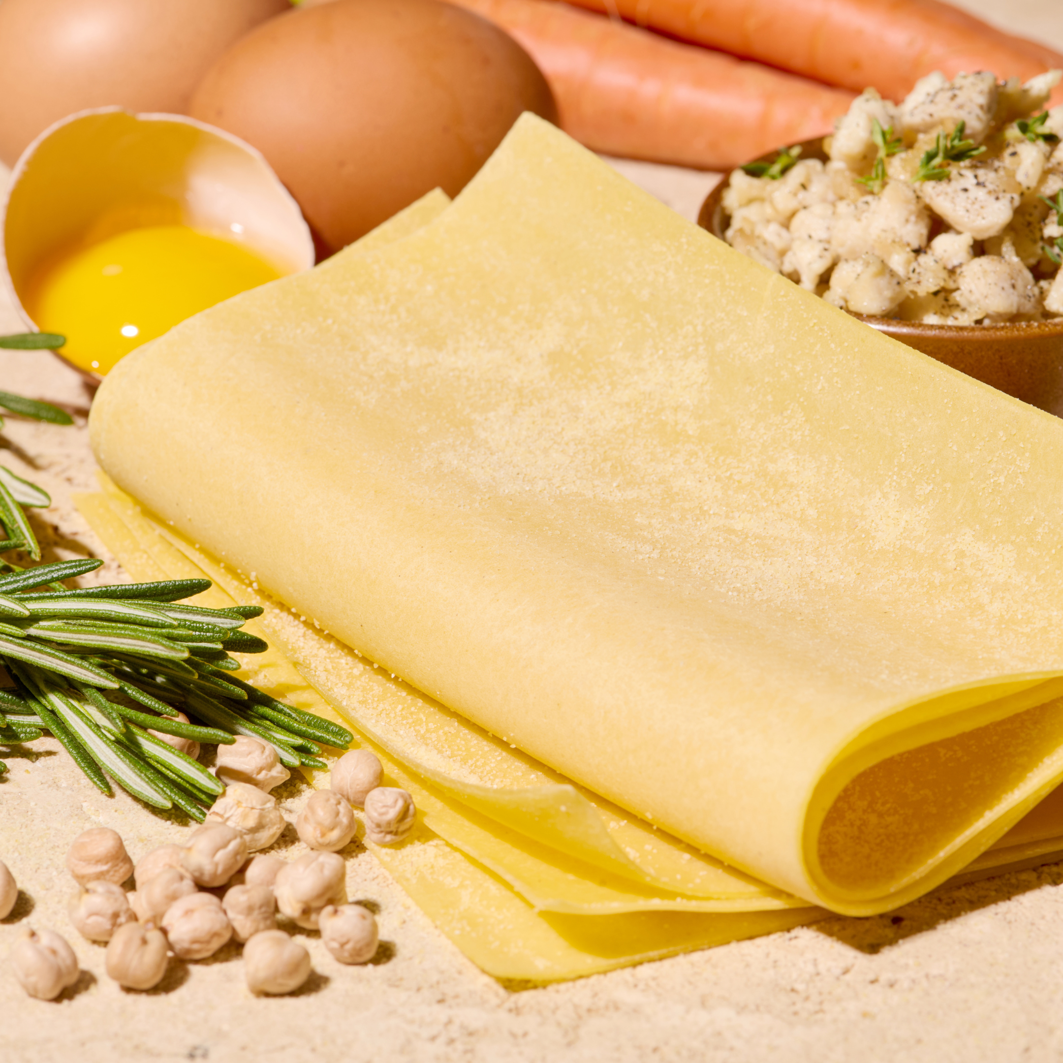 Frische Lasagneblätter mit Kichererbsen, Rosmarin, Karotten und einem aufgeschlagenen Ei auf einem Tisch.