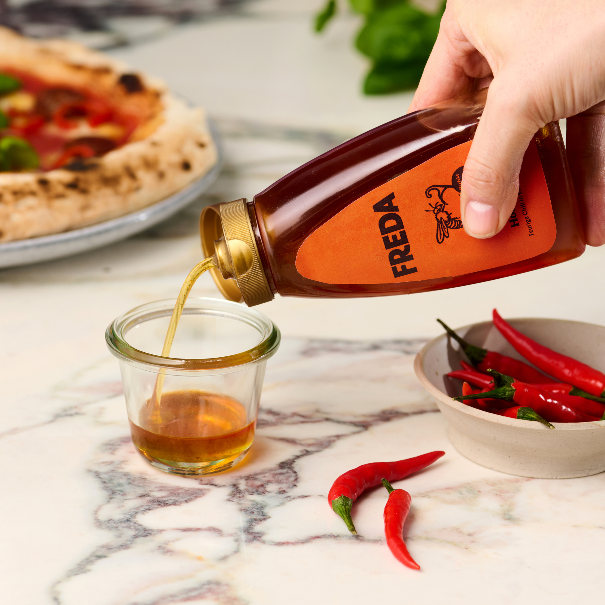 Eine Hand gießt Honig aus einer Flasche in ein Glas vor einem Teller mit roten Chilischoten und einer Pizza.