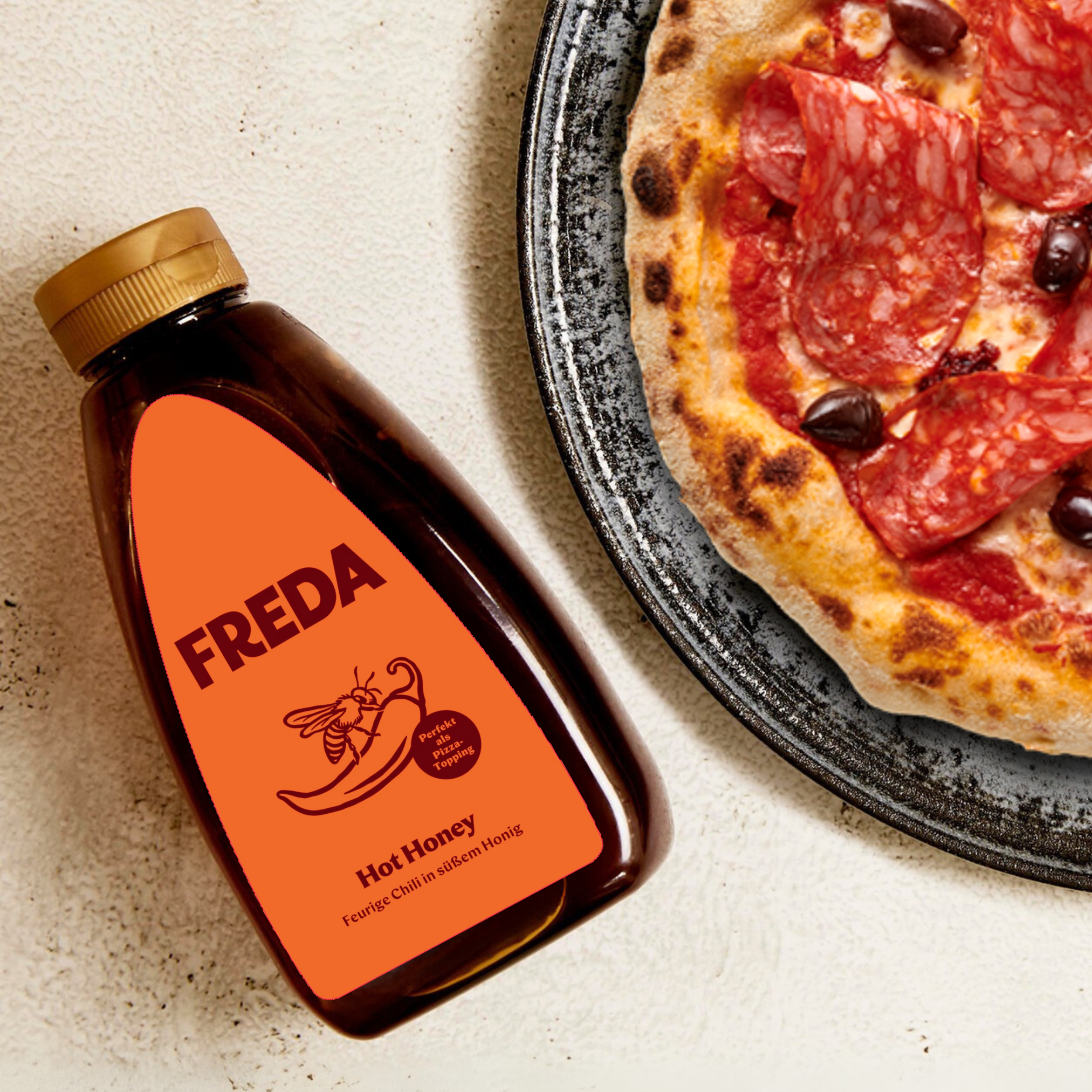 Eine Flasche Freda Hot Honey neben einer Pizza mit Salami und Oliven auf einem Teller.