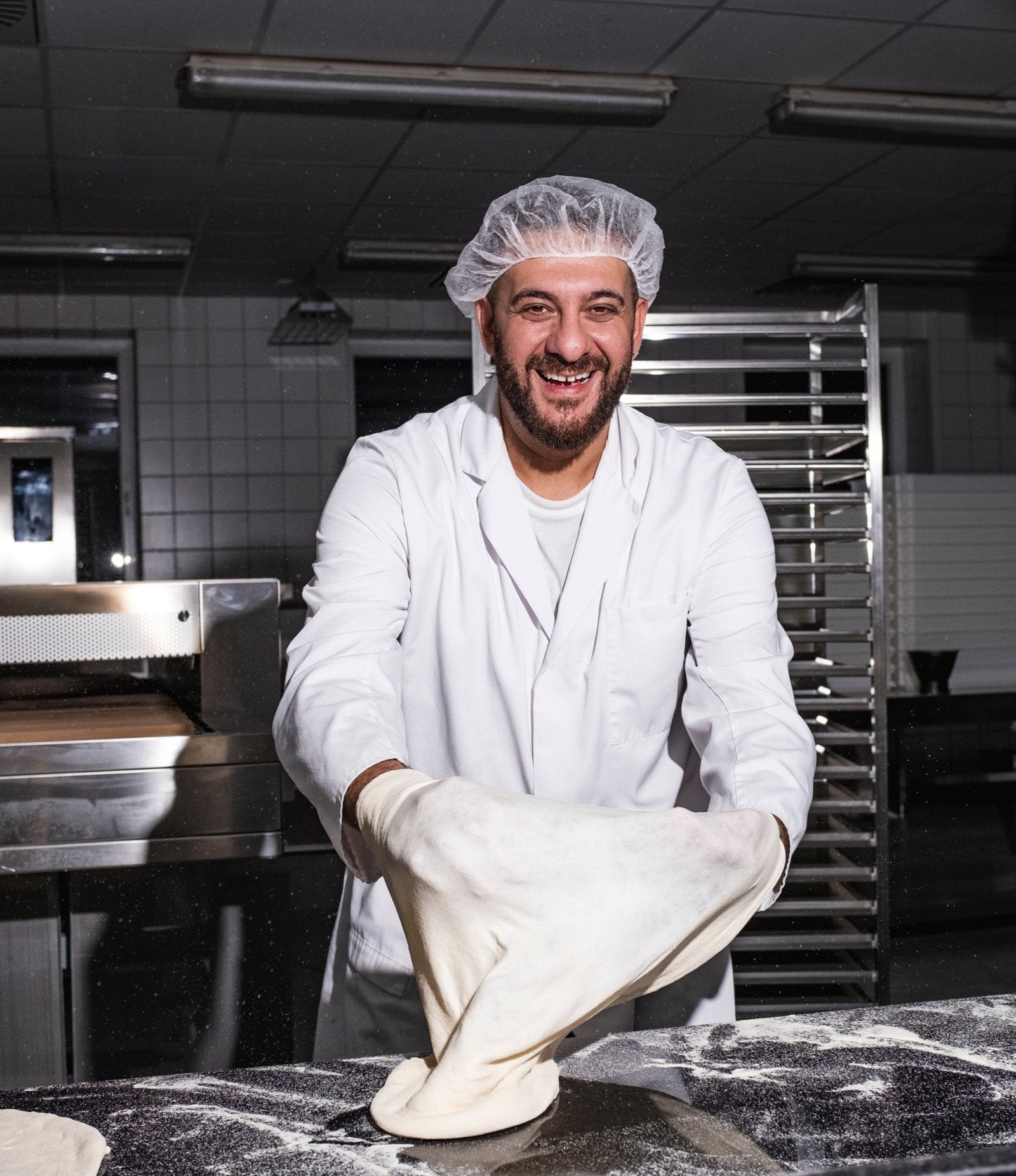 Lächelnder Bäcker mit Schutzhaube und weißem Kittel formt Teig in einer Bäckerei.