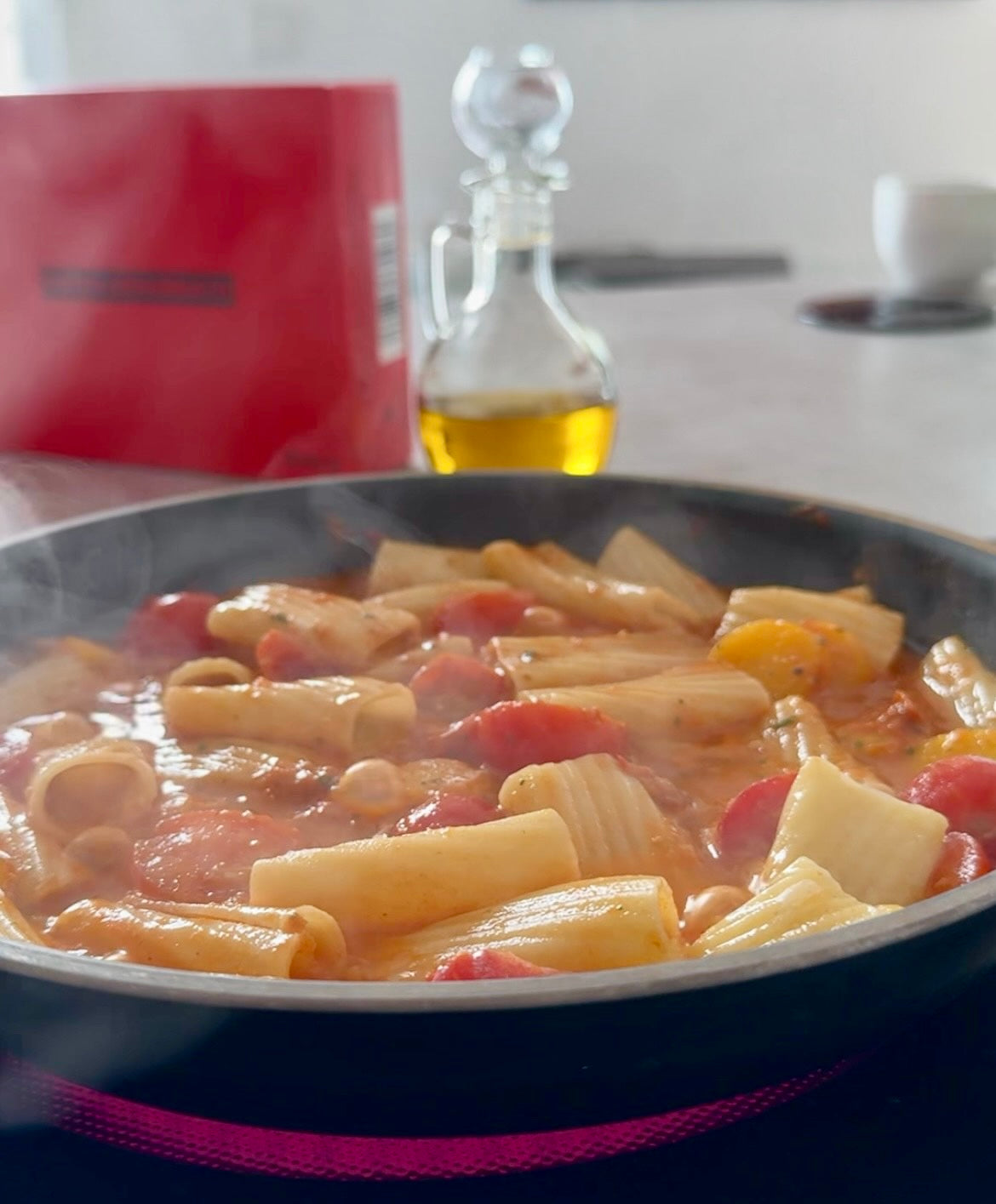 Pfanne mit dampfender Pasta in Tomatensauce, im Hintergrund eine Ölflasche und eine rote Box.