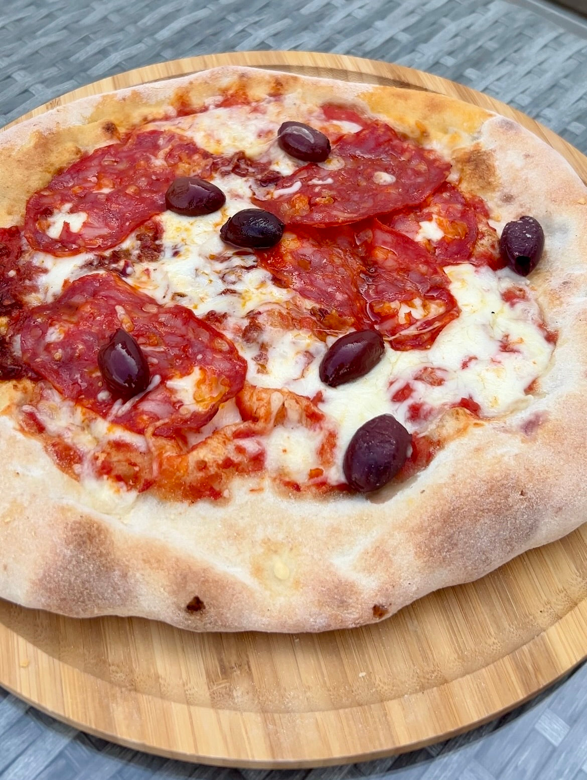 Pizza mit Salami, geschmolzenem Käse und schwarzen Oliven auf einem hölzernen Servierbrett.