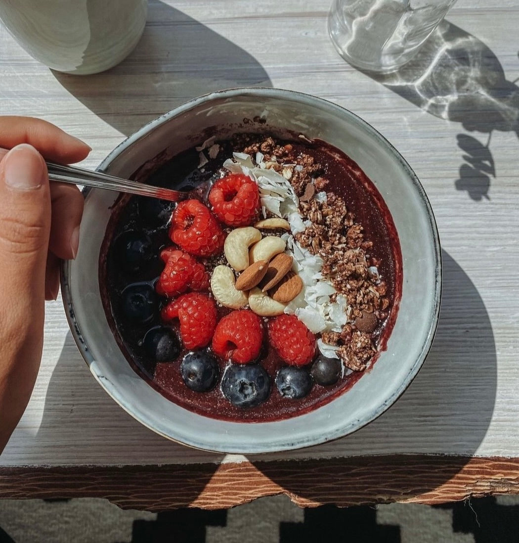 Eine Schüssel Acai-Bowl mit Himbeeren, Blaubeeren, Nüssen und Müsli auf einem Tisch, neben einer Hand mit Löffel.