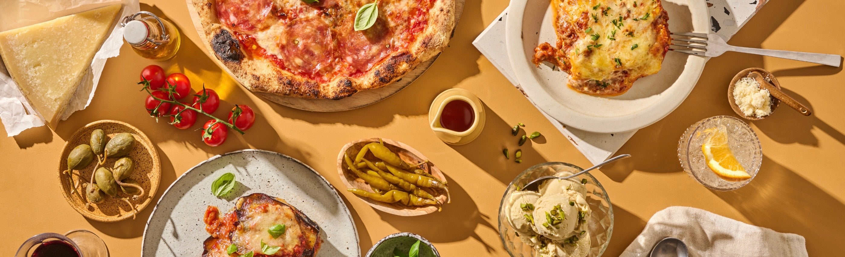 Verschiedene italienische Speisen auf einem Tisch, darunter Pizza, Lasagne, Käse, Kirschtomaten und ein Glas mit Getränk.