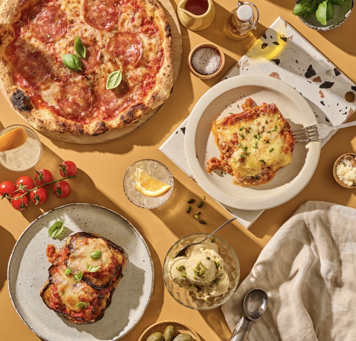 Pizza, Lasagne und Auberginenauflauf auf einem Tisch mit Getränken, Kirschtomaten, Pistazieneis und Gewürzen.