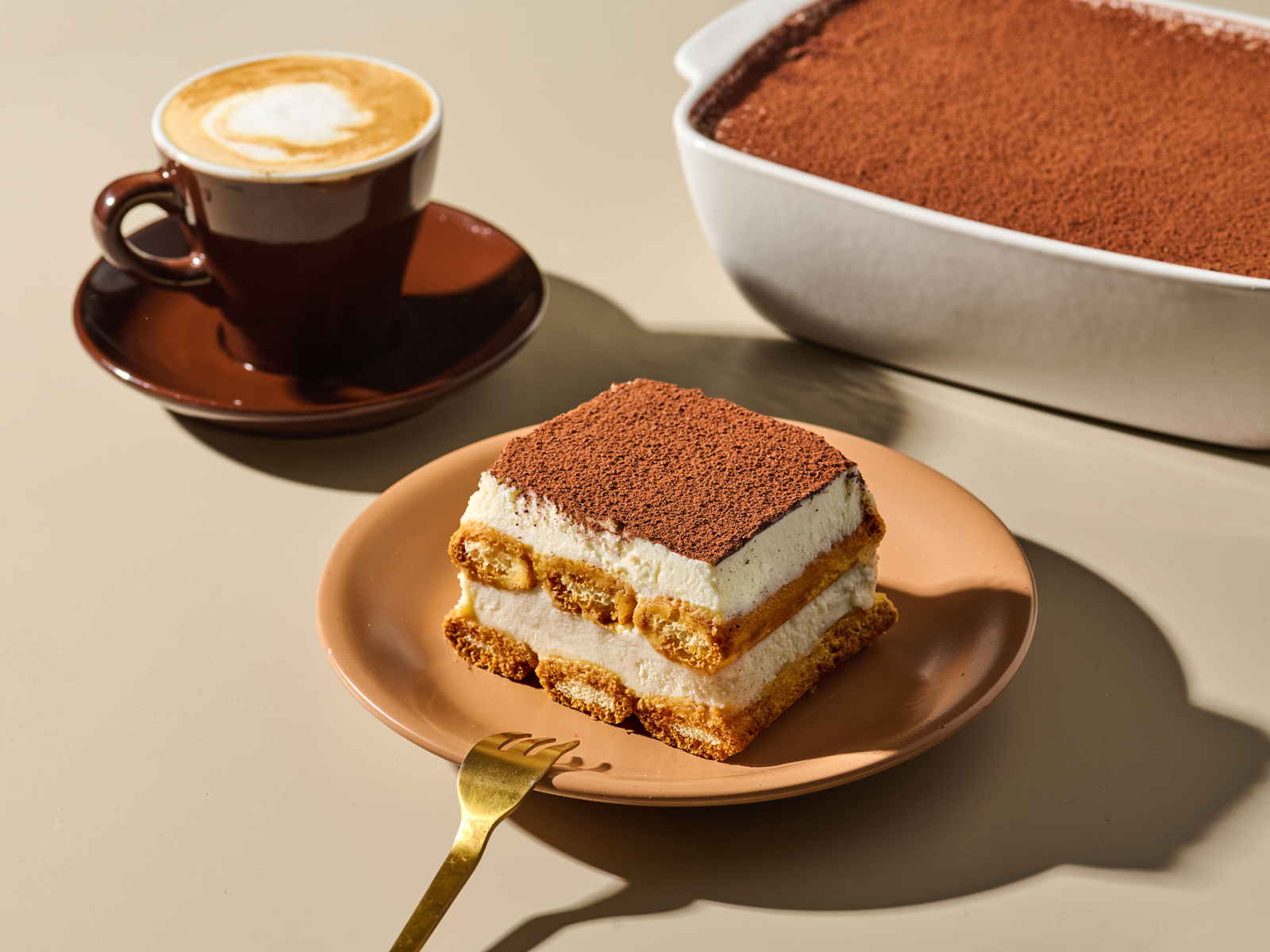 Ein Stück Tiramisu auf einem Teller mit einer Gabel und einer Tasse Kaffee daneben, im Hintergrund eine Auflaufform mit Tiramisu.