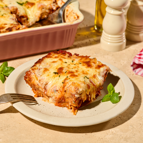 Ein Stück Lasagne auf einem Teller mit Basilikumblättern, daneben eine Auflaufform und Gewürze im Hintergrund.