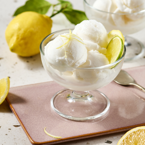 Glas mit Zitronensorbet, garniert mit Zitronenscheiben und Zitronenzesten, auf einem rosa Untersetzer, Zitronen im Hintergrund.