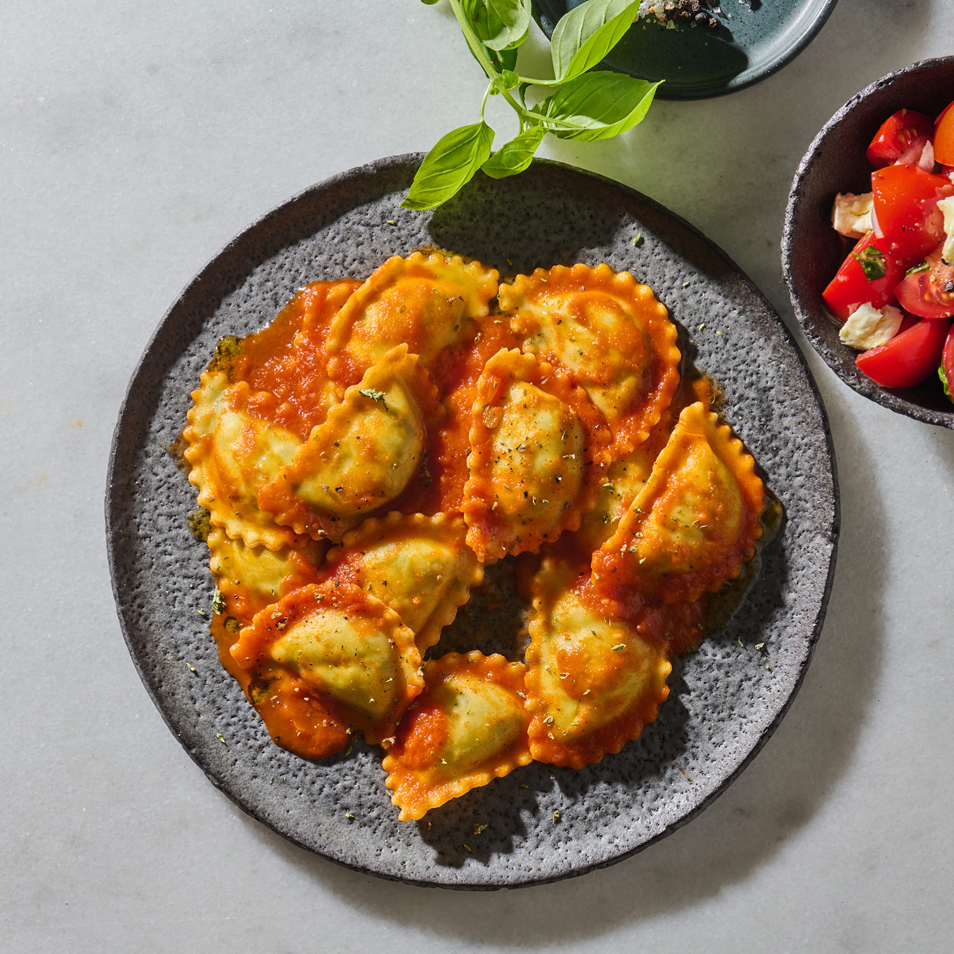 Teller mit Ravioli in Tomatensoße, daneben ein Schälchen mit Tomatensalat und frischen Basilikumblättern.