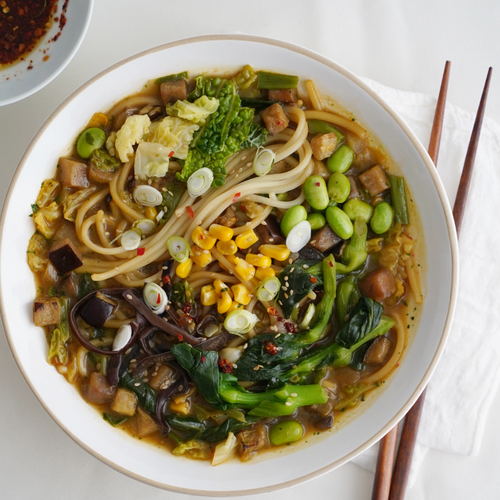Eine Schüssel mit Nudelsuppe, buntem Gemüse und Tofu, daneben Essstäbchen und eine Schale mit Gewürzöl.