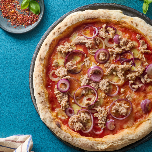 Pizza mit Thunfisch, roten Zwiebeln und Käse auf einem blauen Tisch, daneben ein Teller mit Chiliflocken und Basilikum.