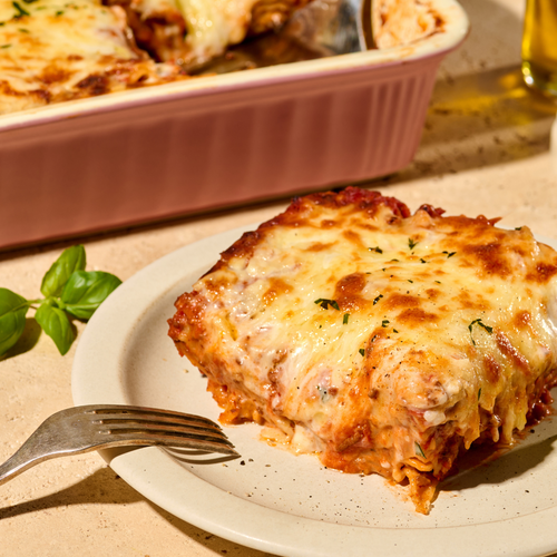 Ein Stück Lasagne auf einem Teller neben einer Gabel, mit einem Basilikumzweig als Dekoration daneben.