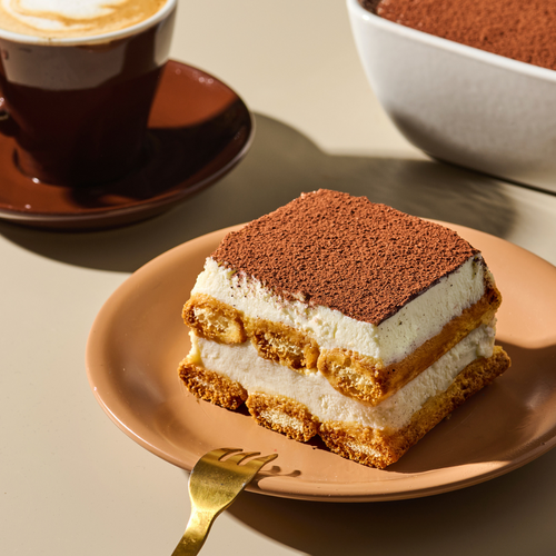 Tiramisu-Stück auf braunem Teller neben einer Tasse Kaffee und einer vollen Schale, im Sonnenlicht auf dem Tisch.