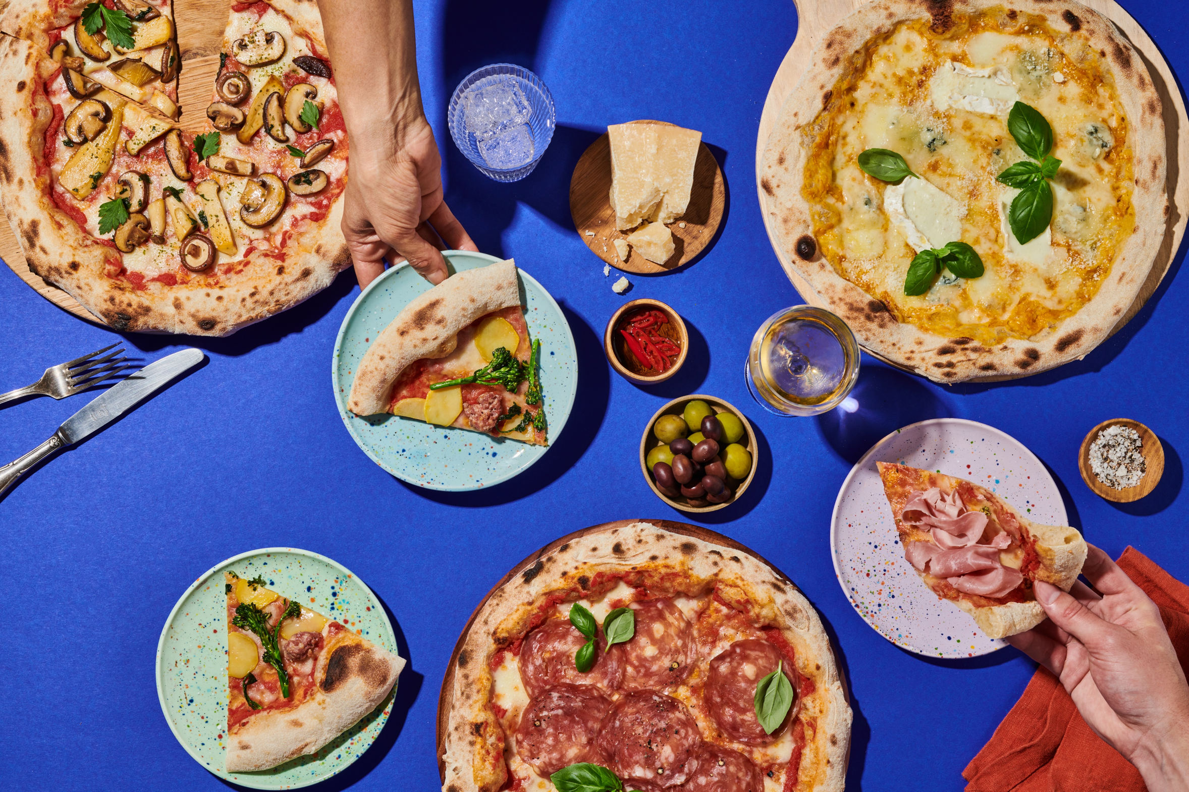 Verschiedene Pizzen und Beilagen auf einem blauen Tisch, mit Händen, die Pizzastücke auf Tellern halten.
