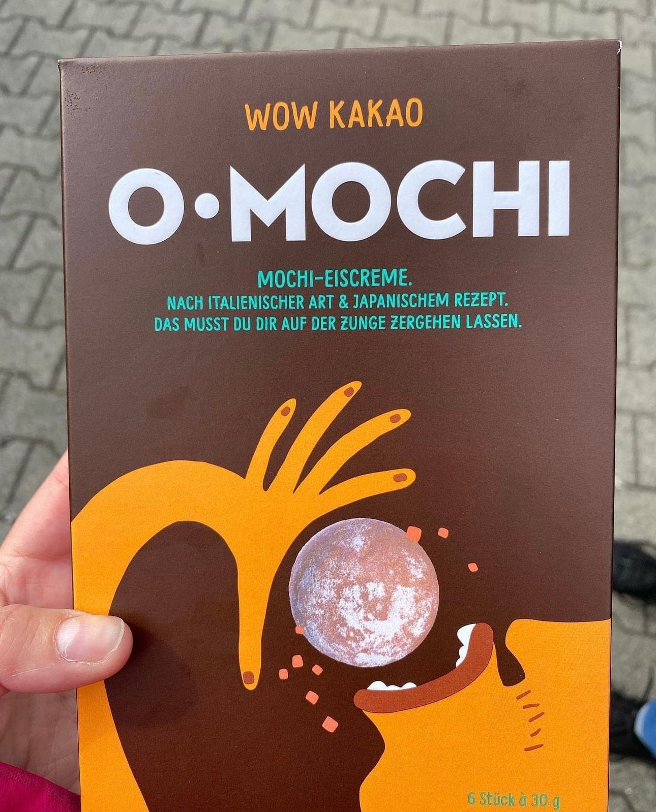 Braune Verpackung von O-Mochi-Kakaoeiscreme, mit illustriertem Gesicht und Hand, die eine Eiscremekugel hält.