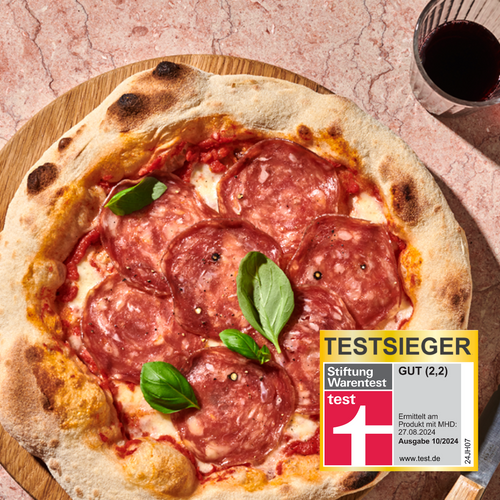 Pizza mit Salami und Basilikum auf Holzbrett mit Testsieger-Etikett, daneben Glas Rotwein.