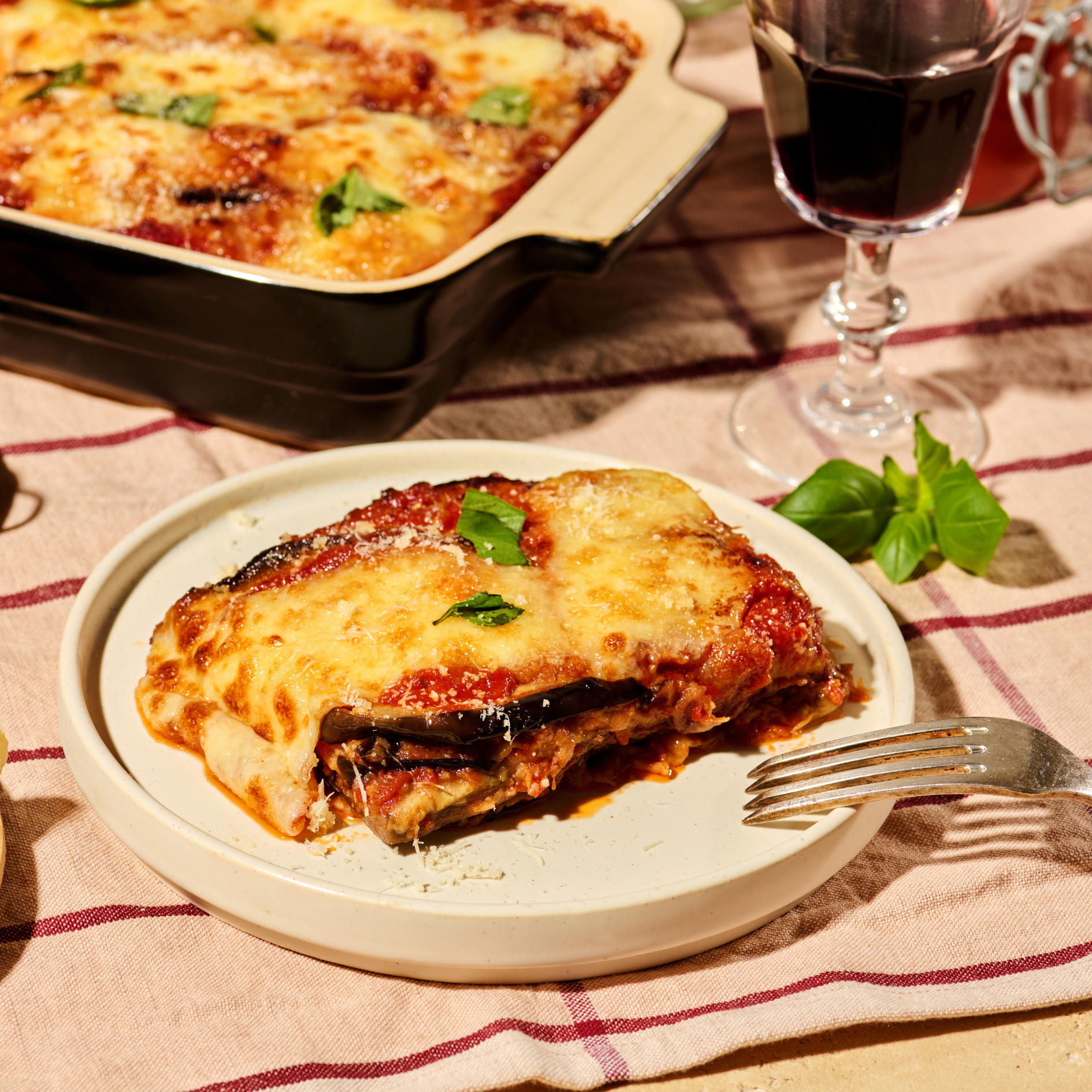 Ein Teller mit Melanzane alla Parmigiana neben einem Glas Rotwein auf einer karierten Tischdecke.