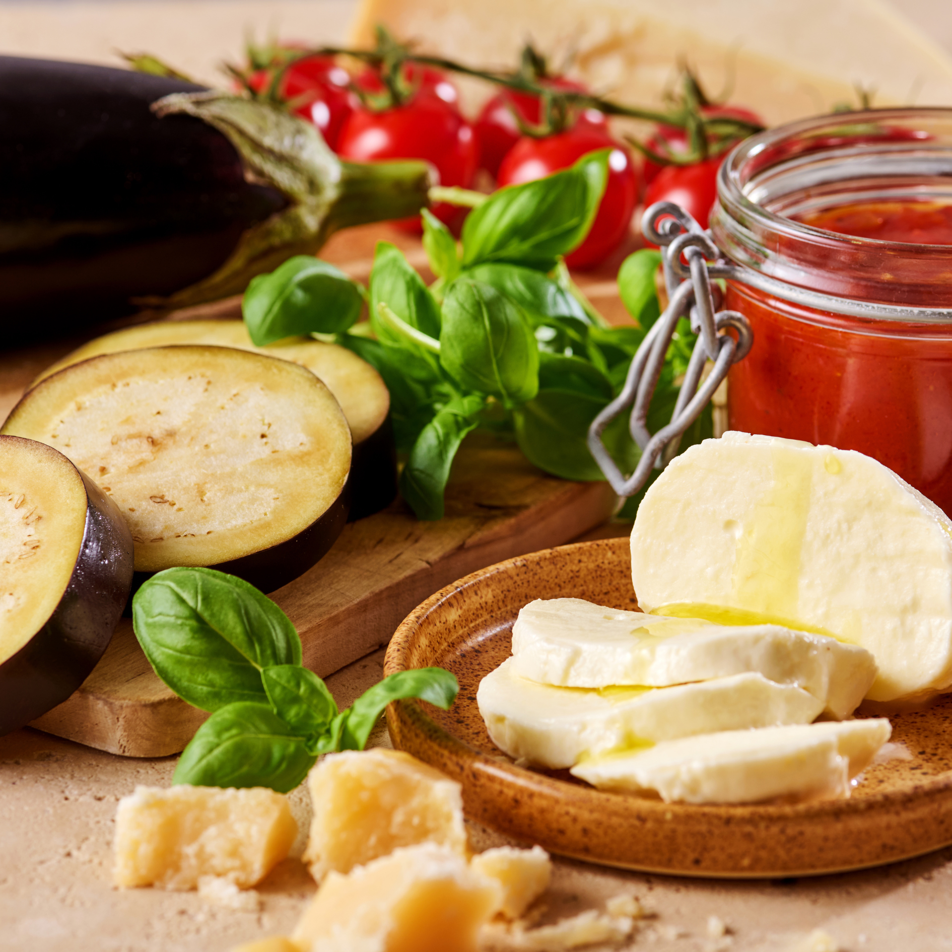 Auberginenscheiben, Mozzarella, Kirschtomaten, Basilikum und ein Glas Tomatensauce auf einem Holztisch.