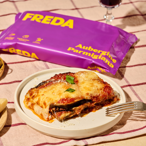 Ein Teller mit Auberginen-Parmigiana steht neben einer lila Verpackung mit gelbem "Freda"-Logo auf einem Tisch.