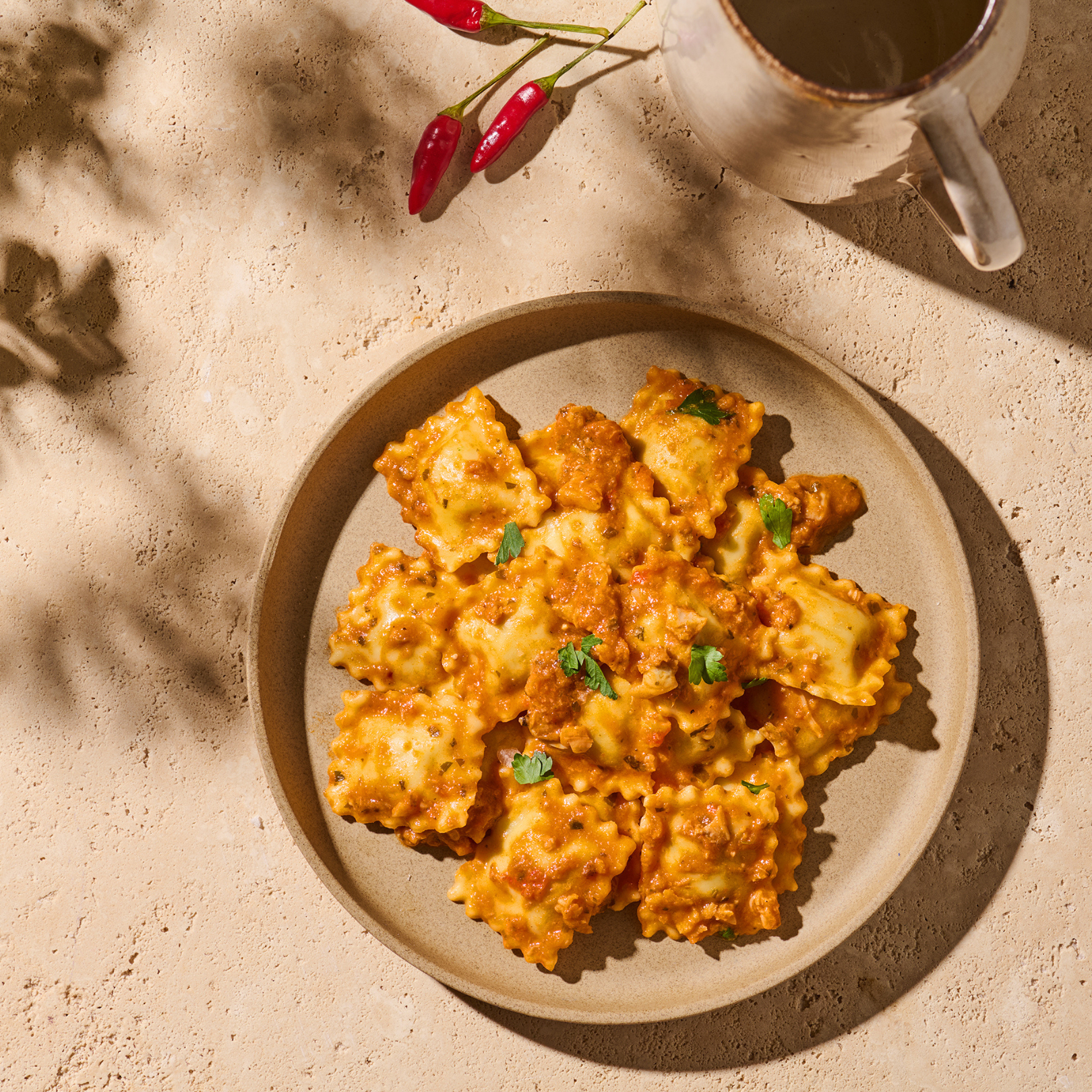 Teller mit Ravioli in Tomatensauce, daneben ein Krug und rote Chilischoten auf sandfarbenem Untergrund.