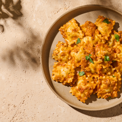 Ein Teller Ravioli in Tomatensauce mit Petersilie garniert, auf einem beigen Untergrund.