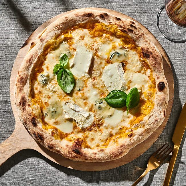 Pizza mit Kürbissauce, Käse und Basilikum auf einem Holzbrett, daneben goldfarbenes Besteck und ein Glas Wein.
