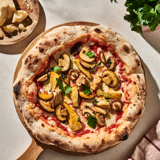 Pizza mit Pilzen und Petersilie auf einem Holzbrett, daneben ein Teller mit Parmesanstücken.