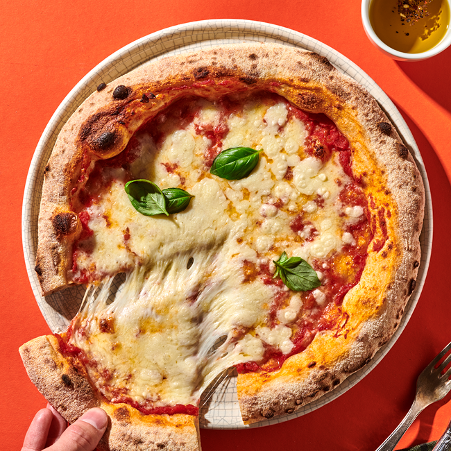 Pizza mit geschmolzenem Käse und Basilikumblättern, eine Hand nimmt ein Stück, daneben eine Tasse Olivenöl.