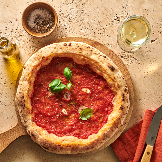 Pizza Marinara mit Basilikum auf Holzbrett, daneben Olivenöl, Gewürze und ein Glas Weißwein auf steinfarbener Oberfläche.